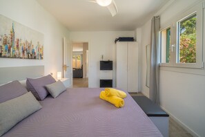 Une chambre Ă coucher comprenant un lit, des oreillers, un ventilateur de plafond, une fenĂȘtre avec des rideaux, un tĂ©lĂ©viseur fixĂ© au mur et une porte donnant sur une autre piĂšce.