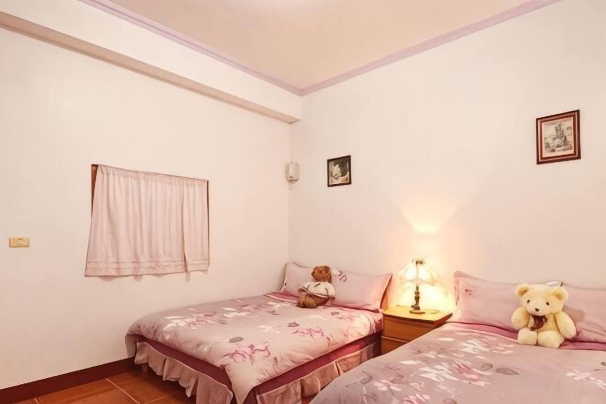 A bedroom with two beds, a nightstand, a lamp, a teddy bear, and a window with curtains.