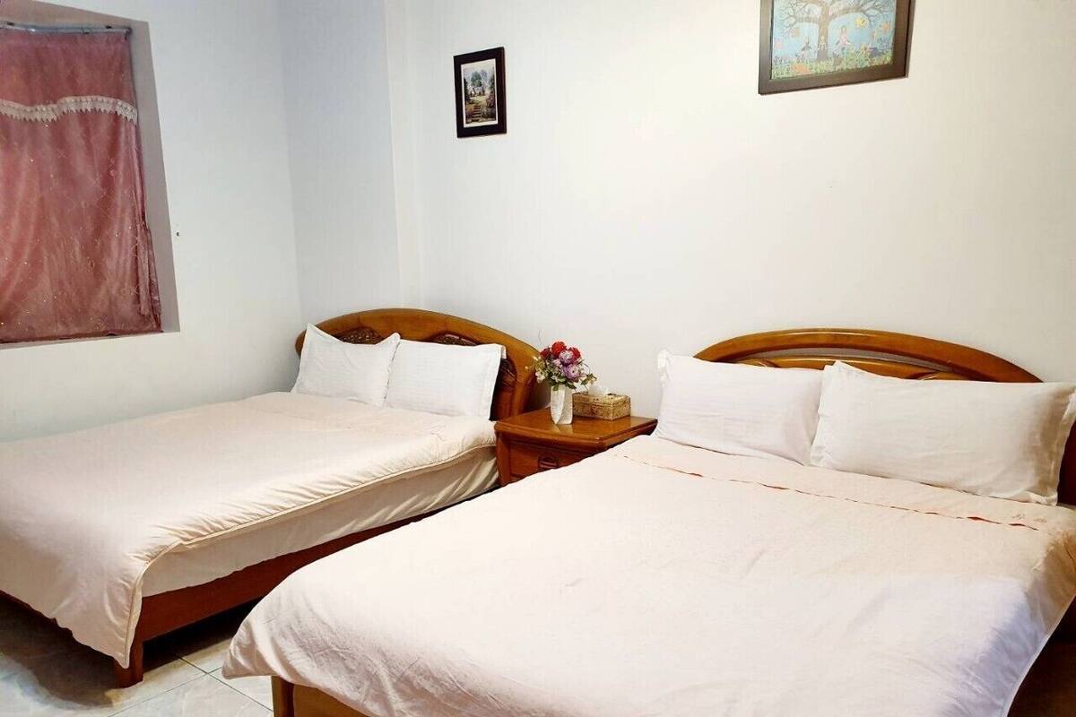 A room with two single beds, wooden headboards, a nightstand with a vase of flowers, and a framed picture on the wall.