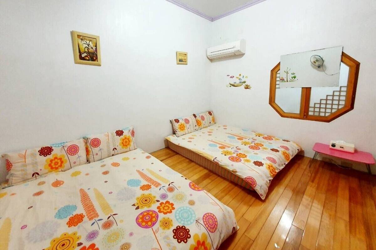 A room with two beds, a wooden floor, an air conditioning unit, a mirror, and a small table with a pink surface.