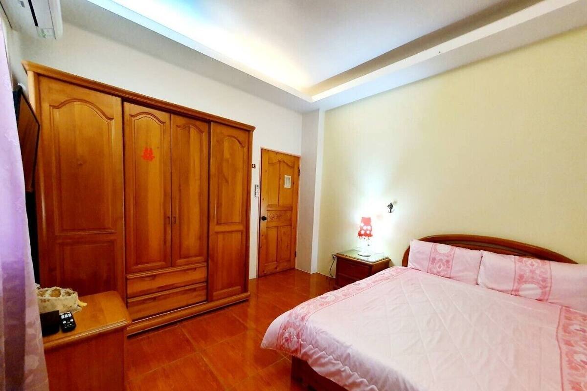 A bedroom with a wooden wardrobe, a bedside table with a lamp, and a bed with pink bedding.