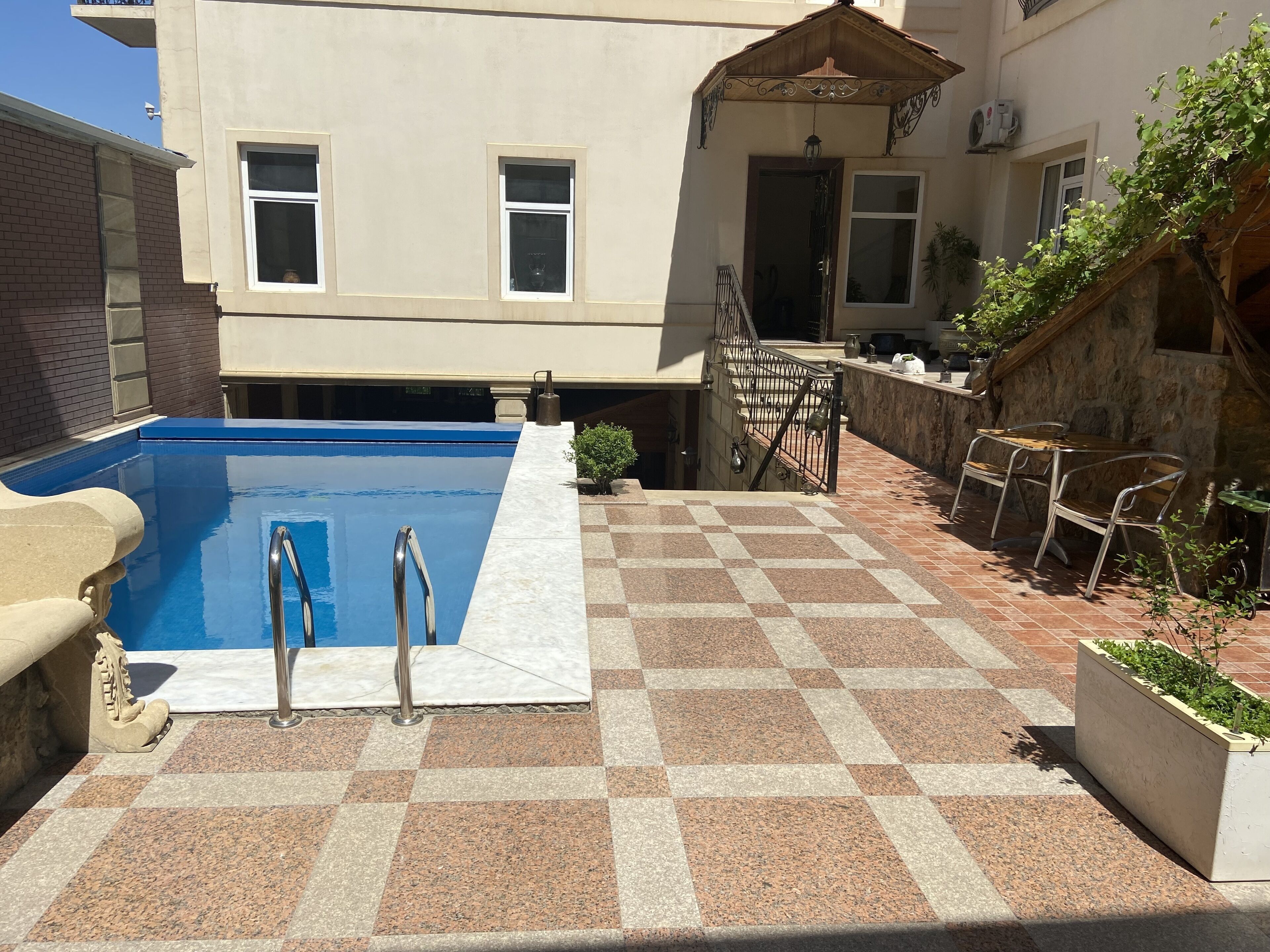 Una piscina con suelo a cuadros, una escalera metálica y un pequeño jardín con mesa y sillas.