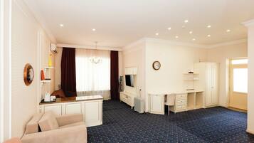 Habitación de hotel con kitchenette, comedor, sala de estar con sofá y televisor, y un ventanal con cortinas.
