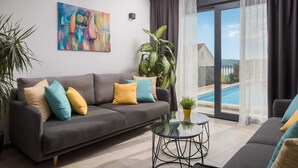 A modern living room with a grey sofa, colorful cushions, a glass coffee table, and a large window with a view of a swimming pool and a building. - Villa Marulovo (Okrug)
