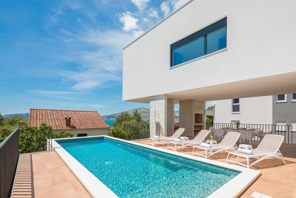 A modern pool area with clear blue water, white lounge chairs, and a view of the sea and mountains in the background.