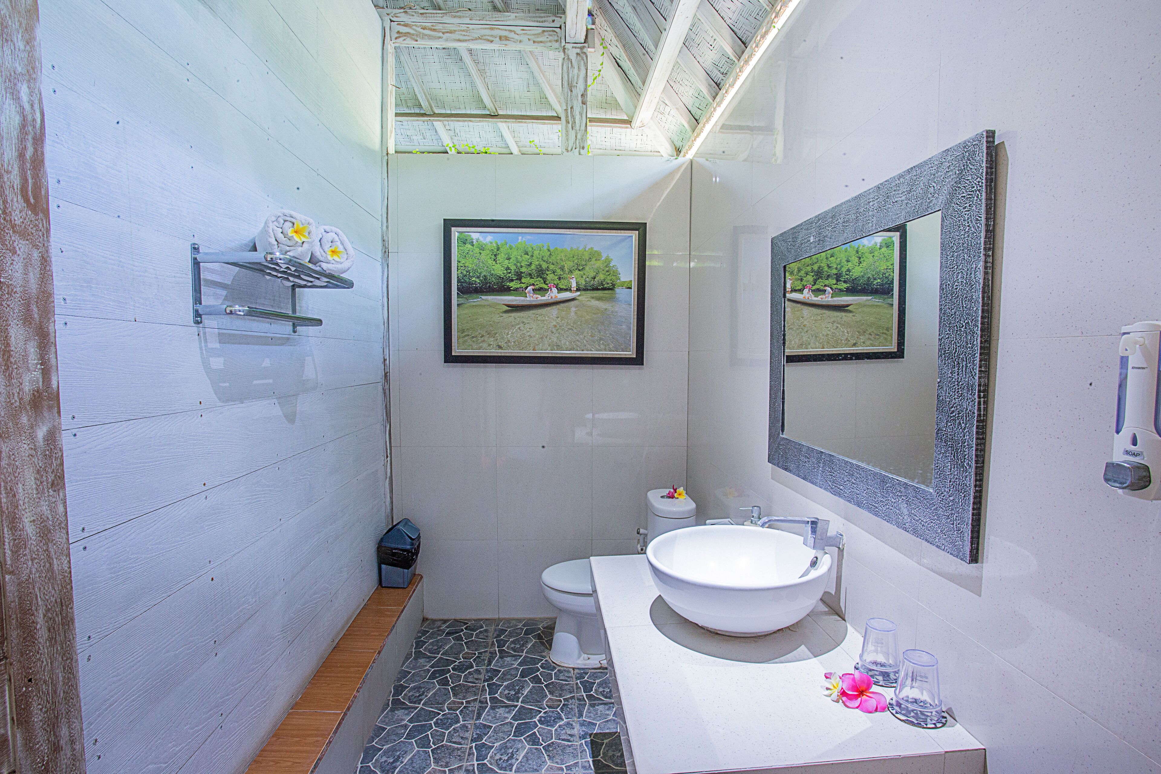 Een badkamer met een witte wastafel, toilet en douche. Er hangen twee afbeeldingen in lijsten aan de muur, één van een boot op een rivier en de ander van een tuin.