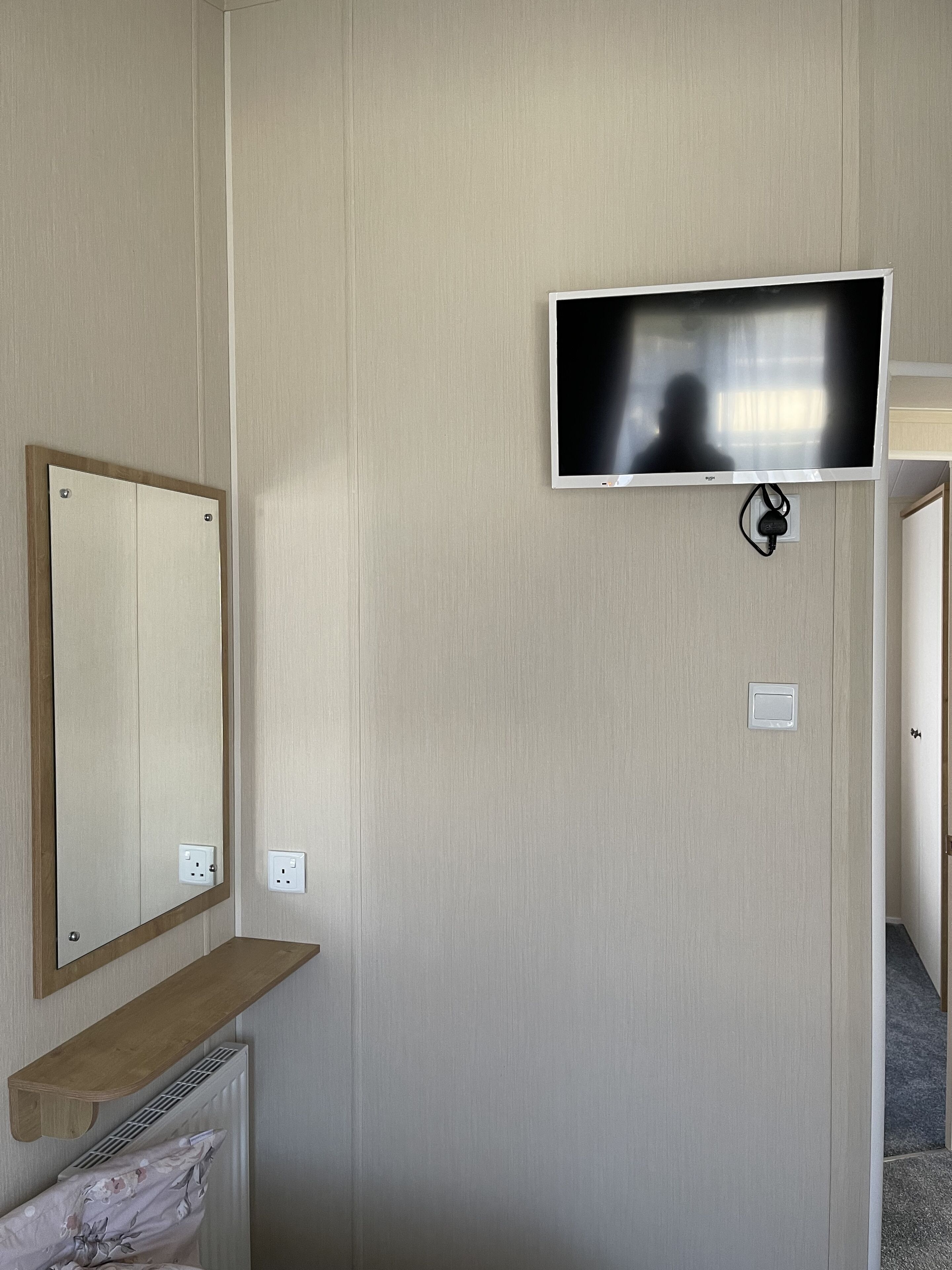 Ein Zimmer mit einem an der Wand befestigten Flachbildfernseher, einem Holzregal, einem Spiegel und einer Steckdose.