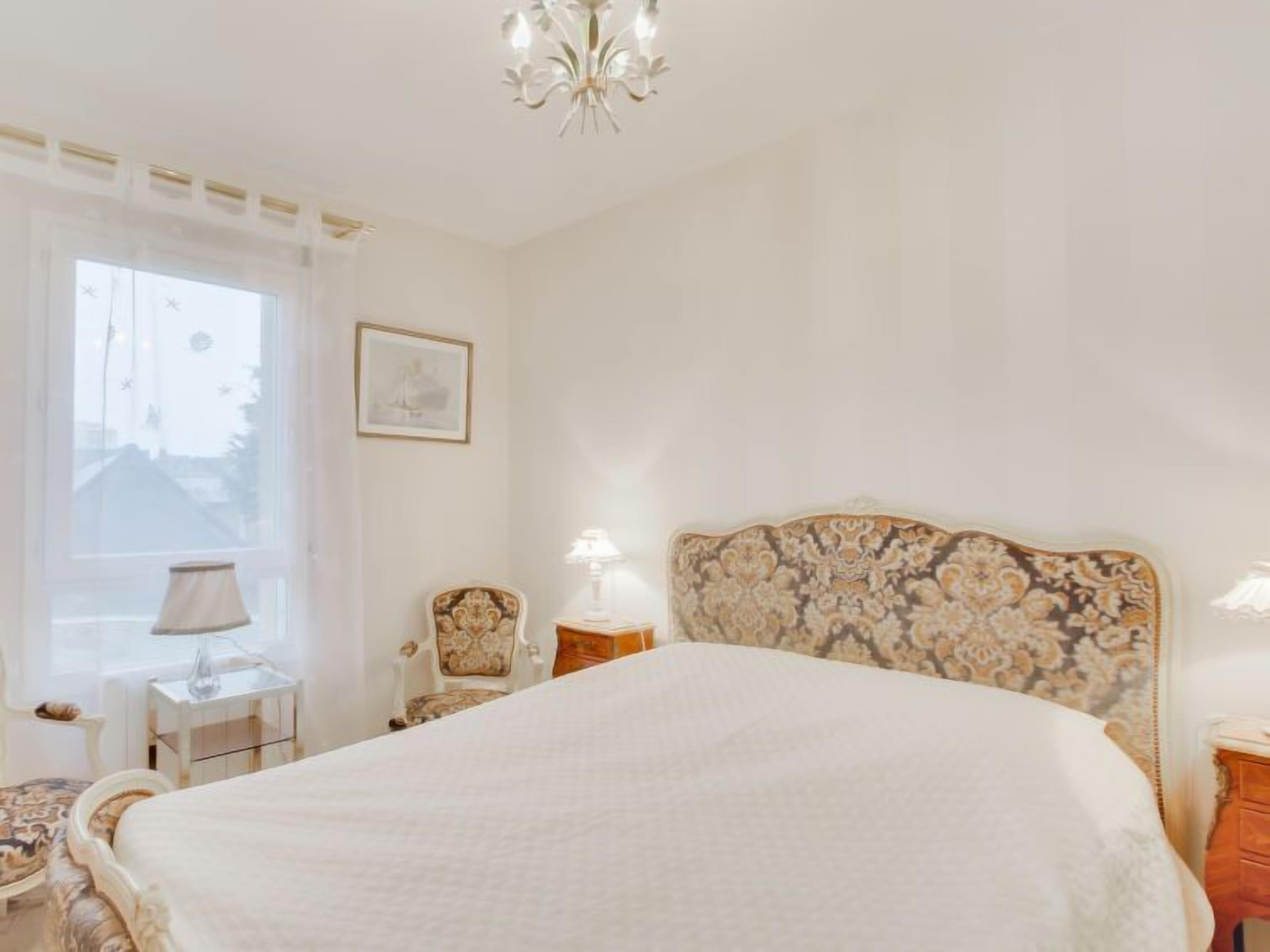 A bedroom with a large bed, a chandelier, a chair, a nightstand, and a window with curtains.