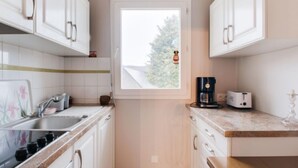 A compact kitchen with white cabinets, a window with a view of trees, a coffee maker, and a toaster on the countertop. - Le Hoche by Interhome (Dinard)