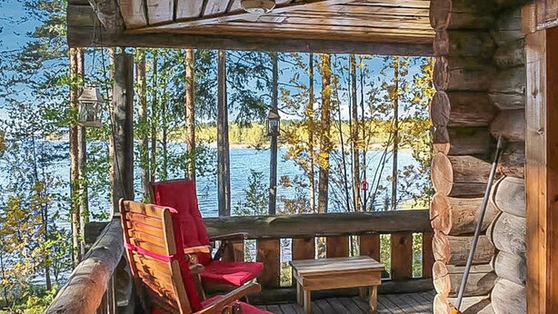 Puinen terassi, jossa on punainen tuoli ja pieni pöytä, ja josta avautuu näkymä puita reunustavalle järvelle.