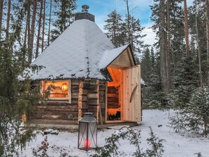 Una cabaña de troncos con techo nevado, interior iluminado y un farol en el exterior.