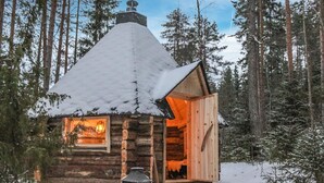 Lumihunnun peittämä hirsimökki, jonka sisätilat ovat valaistuja, ja jonka ympärillä on metsä.