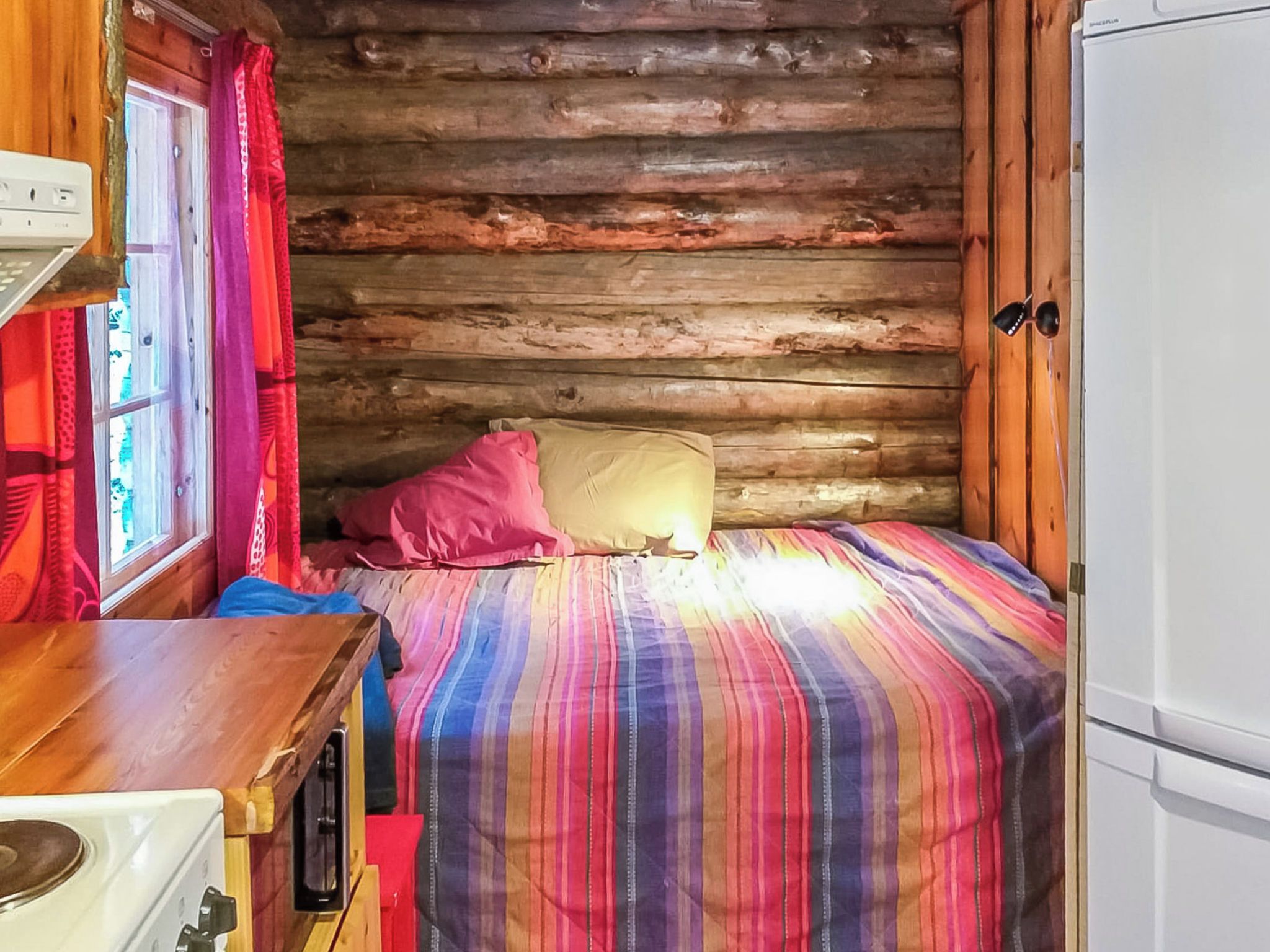 Interior d'una cabana amb una paret de fusta, un llit amb llençols de ratlles, una finestra amb cortines i un electrodomèstic blanc.