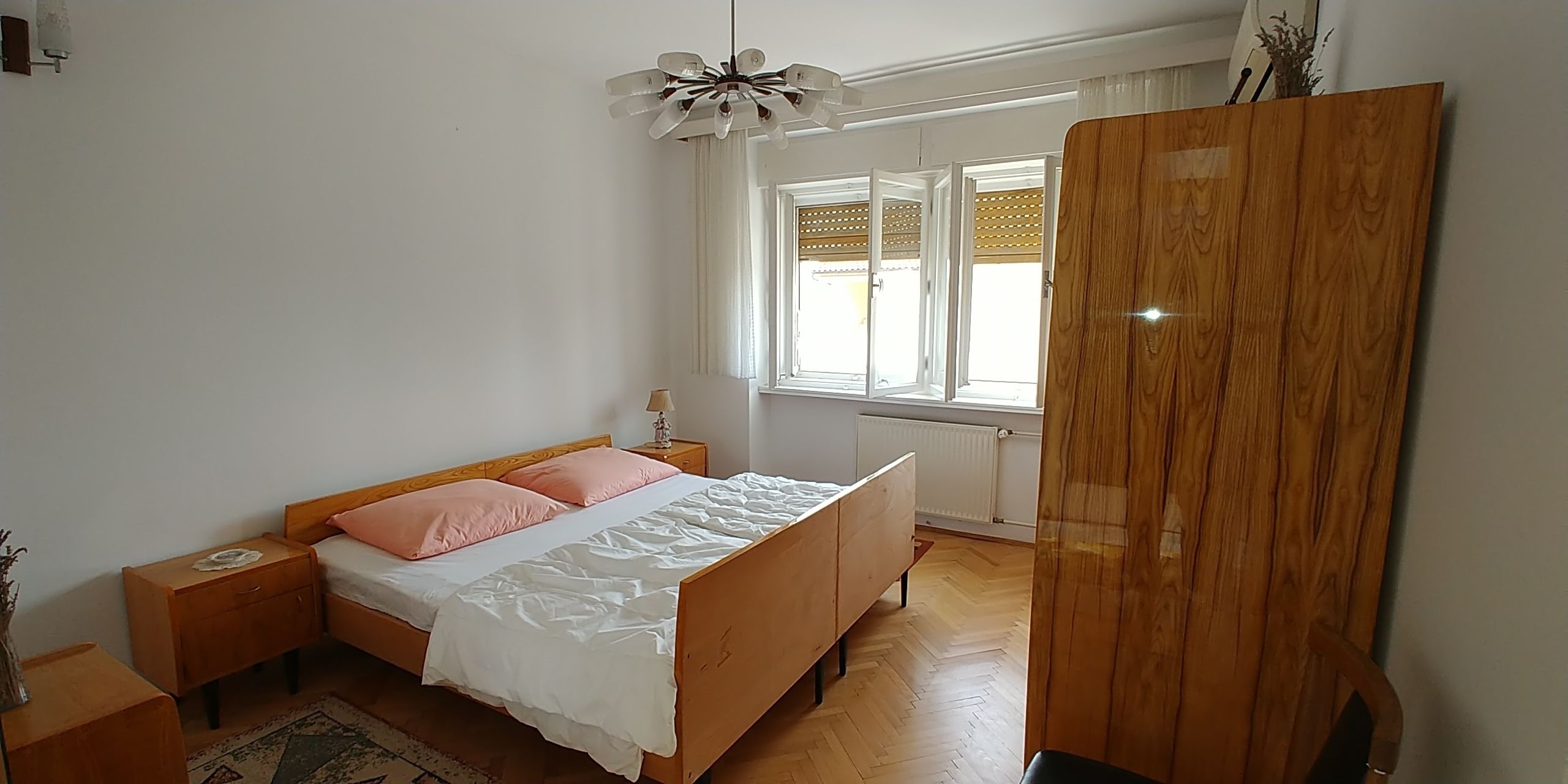 Ein Schlafzimmer mit einem hölzernen Bett, einem Nachttisch, einer Lampe, einem Fenster mit Jalousien und einem Kleiderschrank.