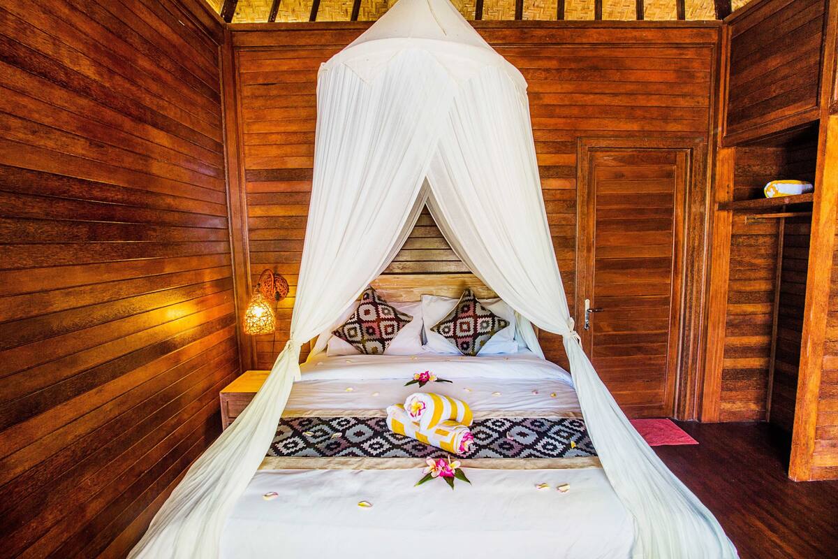 Una habitación de madera con una cama bajo un dosel, almohadas con diseños geométricos y una mesita de noche con una lámpara.