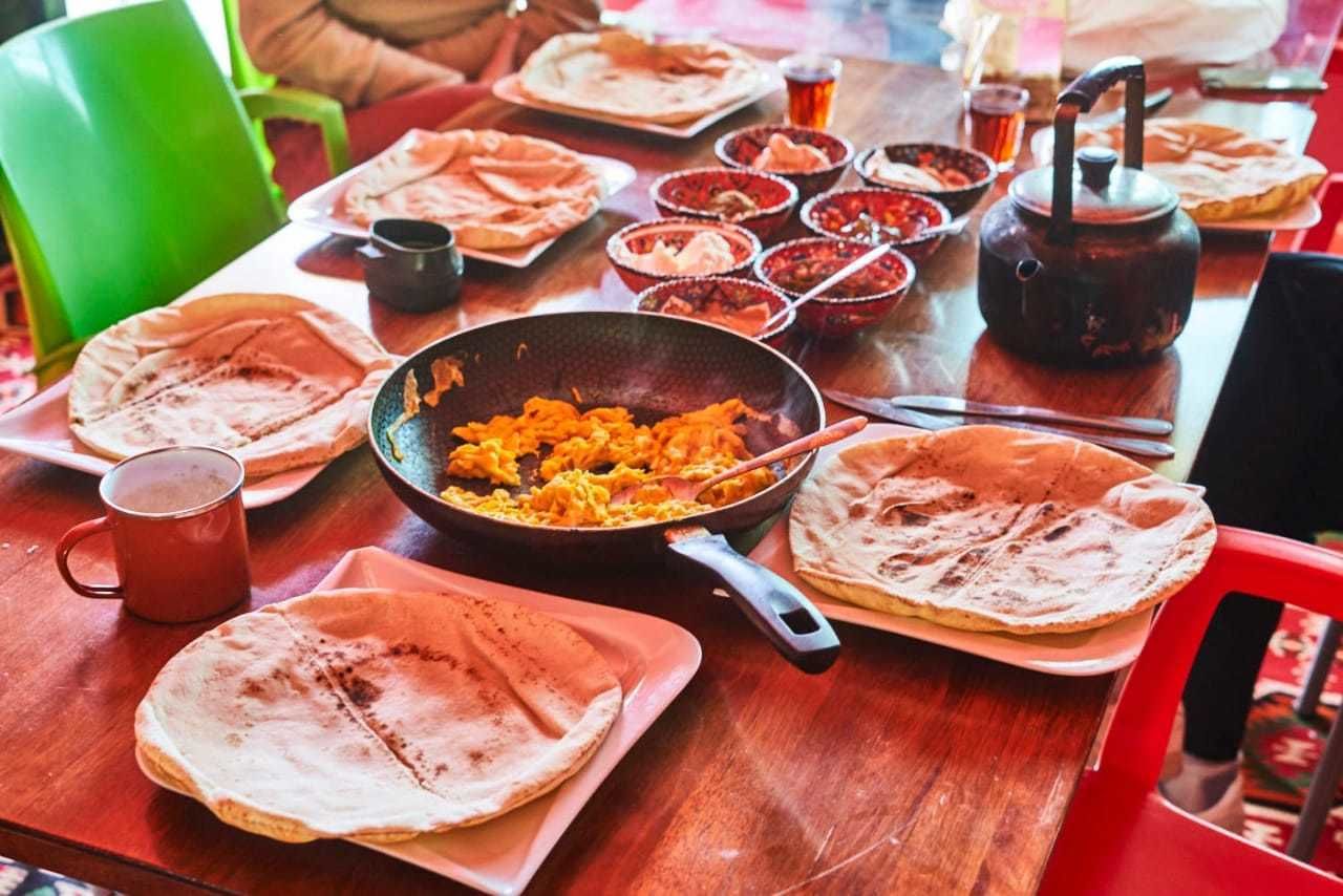 A wooden table with flatbreads, a pan of food, bowls of sauces, and a teapot.