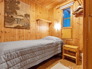 A wooden room with a bed, a small wooden nightstand, a wall-mounted lamp, and a window with a blue blind.