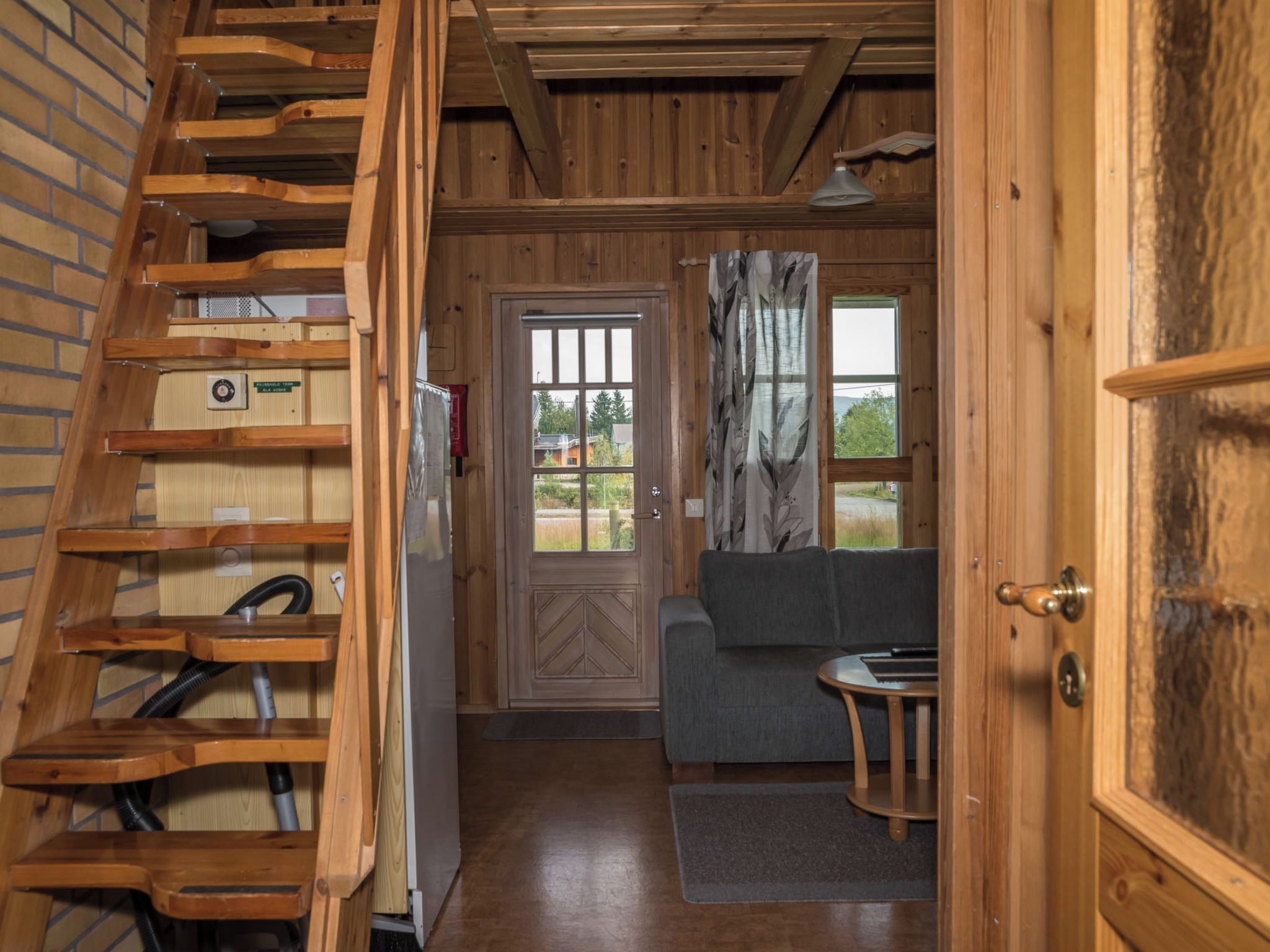 A wooden interior with a staircase, a door leading to a room with a gray sofa, and a window with a view.