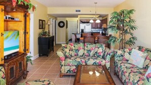A living room with floral-patterned sofas, a wooden coffee table, and a television on a cabinet. - Two-bedroom condo with beautiful Gulf & pool view, just steps from the beach! (Miramar Beach)