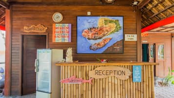 Uma mesa de recepção com uma placa de madeira que diz "RECEPTION" e uma placa de "BEM-VINDO" com um desenho de peixe. Há um relógio, um mapa e uma placa indicando "FREE WIFI".
