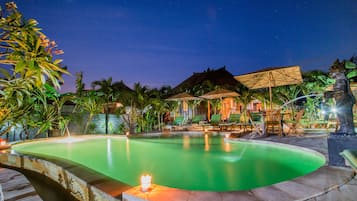 Uma área de piscina à noite, com espreguiçadeiras e guarda-sóis, cercada por plantas tropicais.