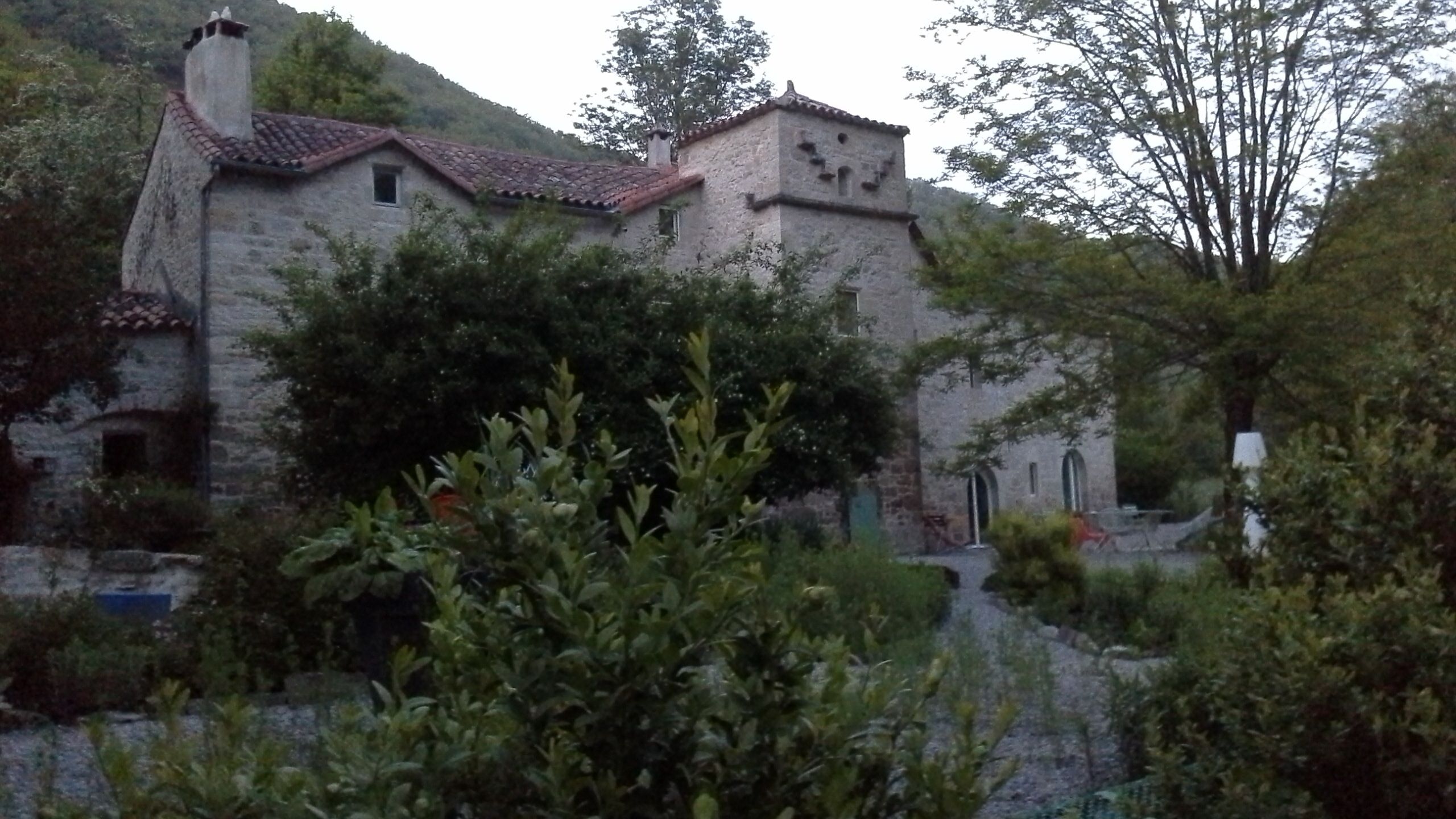 Un bâtiment en pierre, doté d’un toit en tuiles, entouré de verdure et bordé d’un sentier.