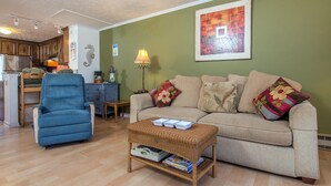 A living room with a blue armchair, a beige sofa with floral cushions, a wicker coffee table, and a kitchen in the background.
