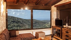 Une pièce dotée d’une grande fenêtre offrant une vue panoramique sur les collines, d’un plafond en bois, d’un canapé, d’une table avec des verres et d’un téléviseur posé sur un meuble en bois.