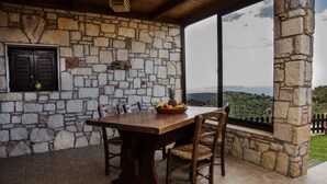Un espace repas agrémenté d’un mur en pierre, avec une table et des chaises en bois, une fenêtre donnant sur le paysage et un panier de fruits posé sur la table.