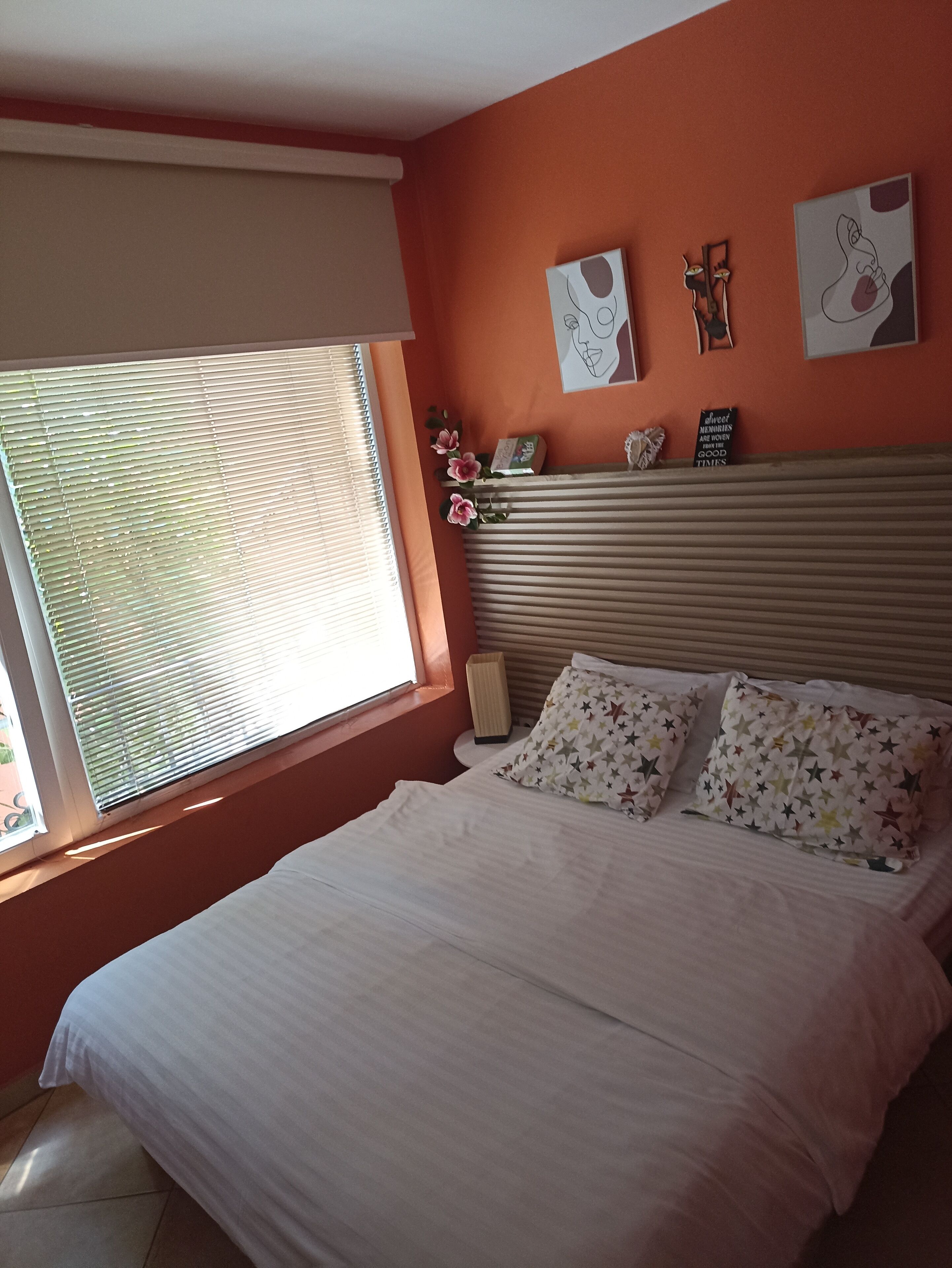 A bedroom with a bed, a window with blinds, and artwork on the wall.