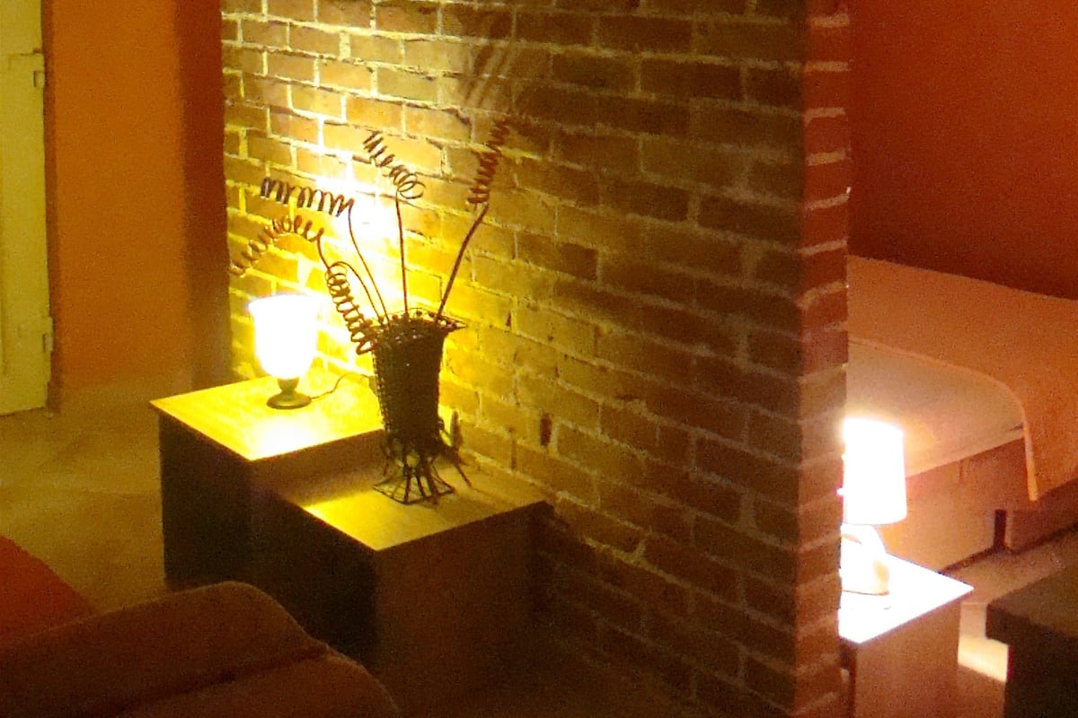 A room with a brick wall, a bed, a small table with a lamp, and a wall-mounted air conditioning unit.