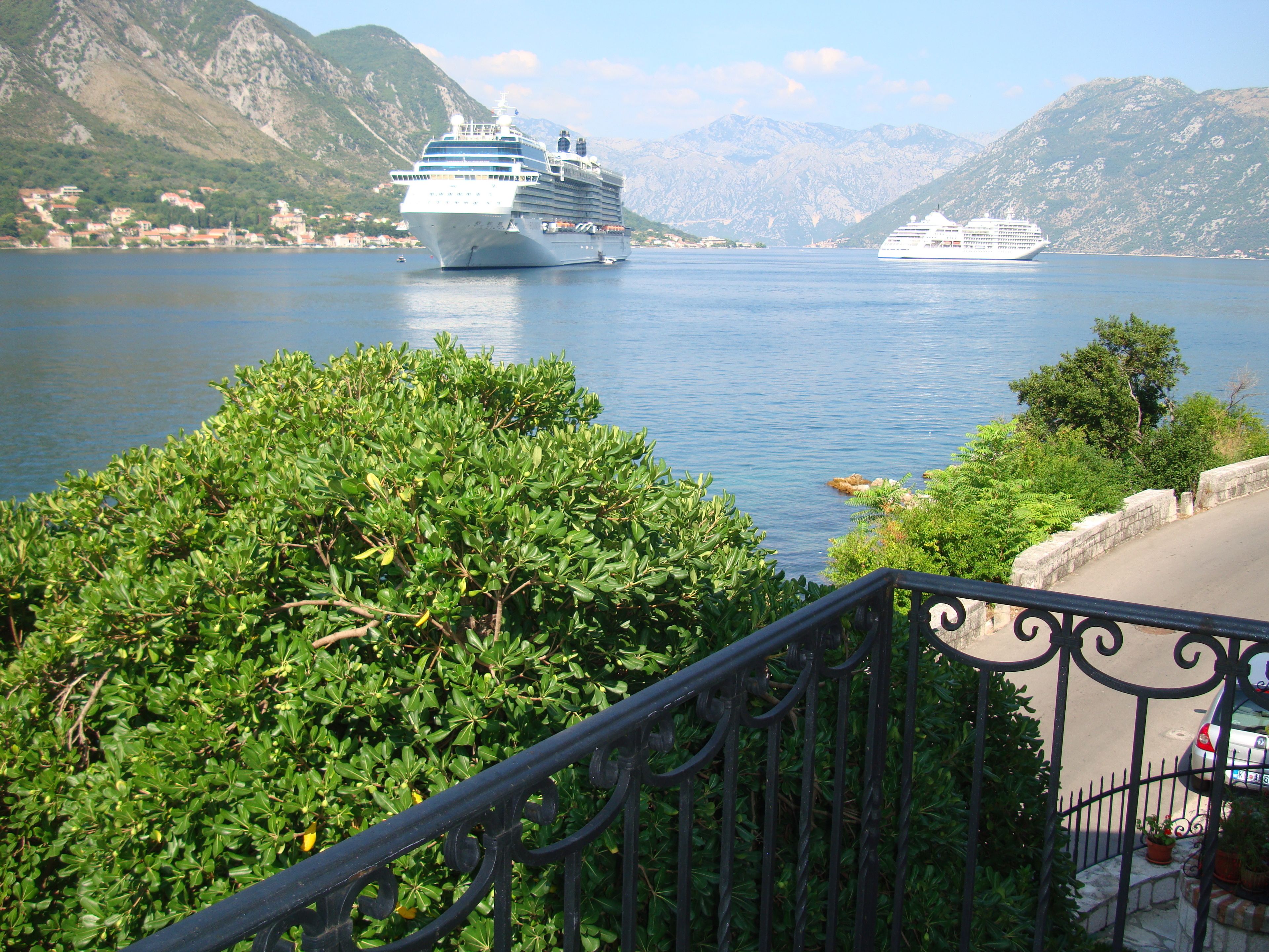 A view of a body of water with two cruise ships, a mountainous landscape, and a balcony with a railing.
