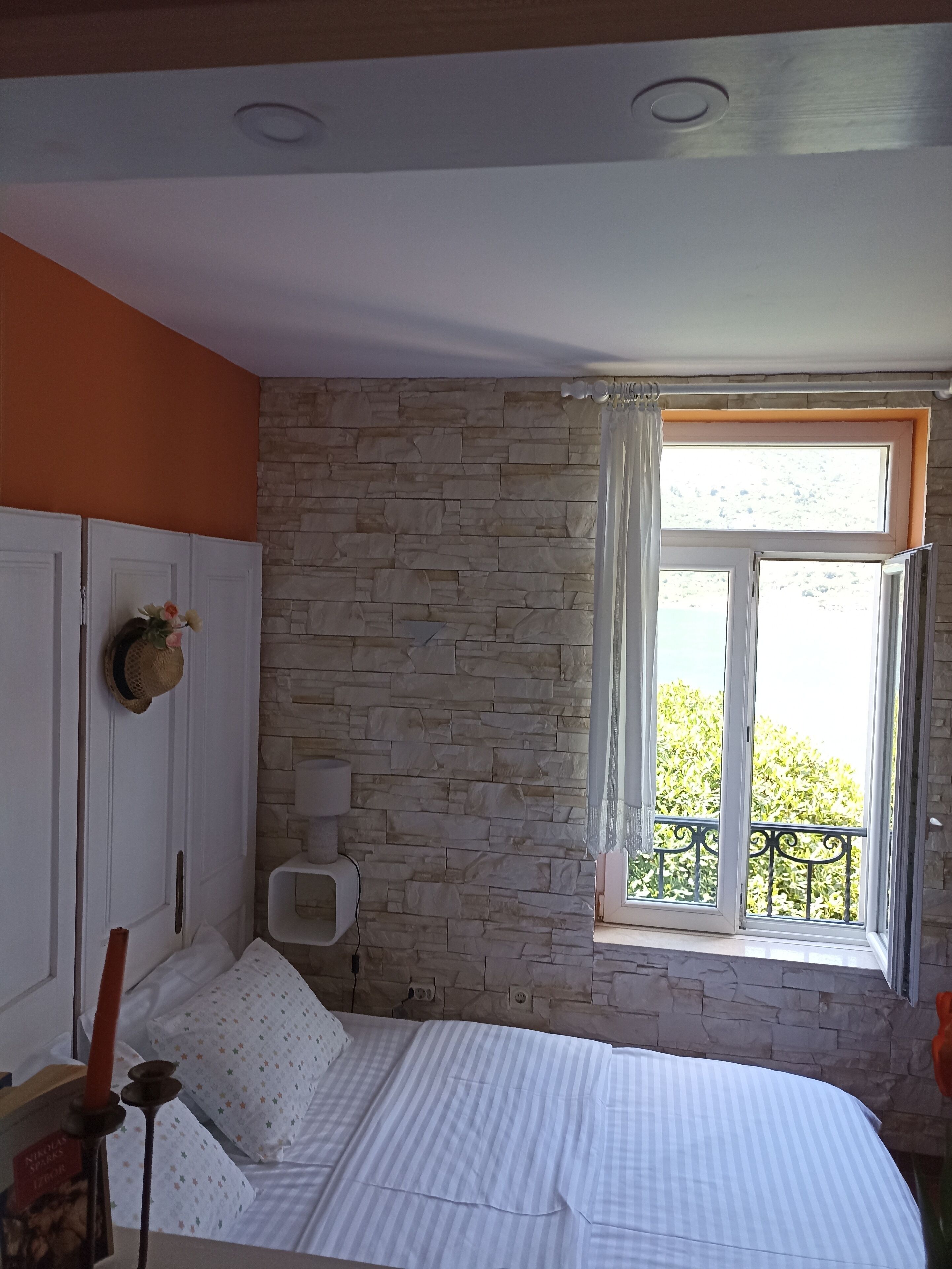 A bedroom with a stone wall, a window with a view of greenery, a bedside table, and a bed with striped bedding.