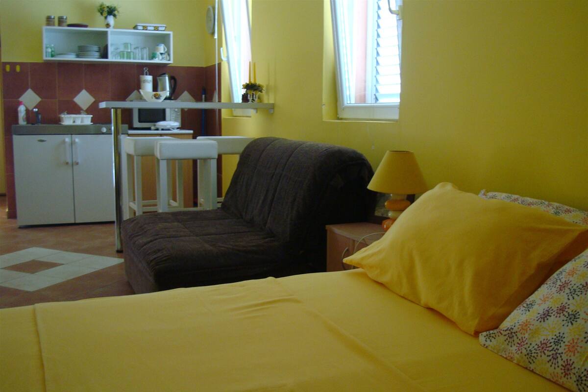 A room with a bed, a chair, a desk, and a kitchenette.