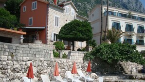 Un hotel frente al mar con paredes de piedra, sillones blancos con sombrillas rojas y agua azul cristalina.