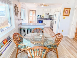 A dining area with a glass table and wicker chairs, a kitchen in the background, and a view through a window. - Inlet Point Villas 4C (North Myrtle Beach)