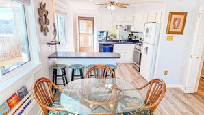A dining area with a glass-top table surrounded by wicker chairs, a kitchen in the background, and a window with blinds. - Inlet Point Villas #4C! Relax & Recharge in Cherry Grove’s Most Peaceful Corner (North Myrtle Beach)
