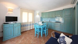 Cocina compacta con armarios color turquesa, comedor con sillas azules, televisor sobre un mueble y ventana con cortinas.