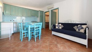 Un espacio habitable compacto con área de cocina que presenta gabinetes color turquesa y una mesa de comedor con sillas azules. Un sofá blanco con cojines estampados se encuentra en la zona de estar.