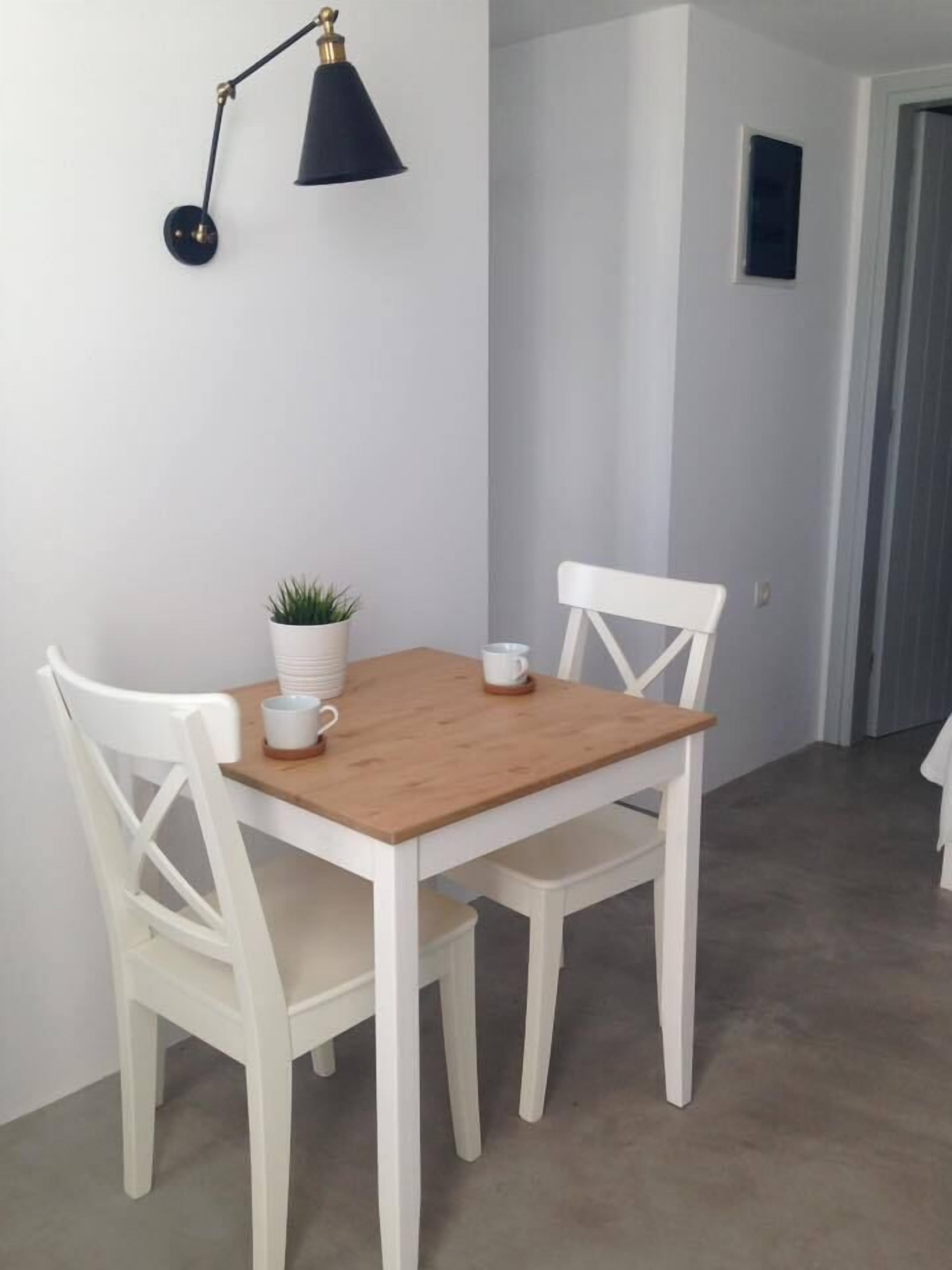 A small dining area with a wooden table and white chairs, a potted plant, and a wall-mounted lamp.