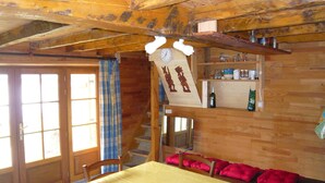 A rustic wooden room with a wooden ceiling, a wooden table with a yellow tablecloth, a chair, and a red cushion. There is a wooden shelf with various items, a clock, and a window with curtains.