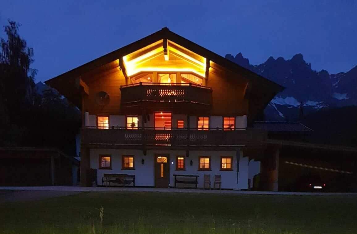 A two-story building with a balcony and multiple windows, surrounded by a grassy area and mountains in the background.