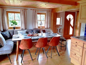 Un comedor con mesa de madera dispuesta para cuatro personas, un sofá y una zona de cocina con armarios de madera.