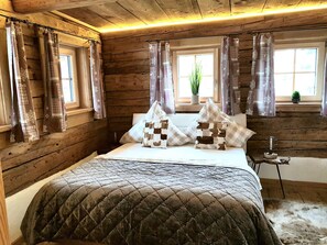 A wooden cabin bedroom with a bed, curtains, and a small table with a lamp.