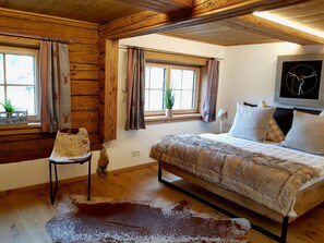 A bedroom with a wooden ceiling, a bed with a fur throw, a chair, a window with curtains, and a plant on the windowsill.