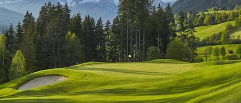 A golf course with a sand bunker, trees, and mountains in the background.