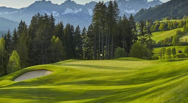 A golf course with a sand bunker, trees, and mountains in the background.