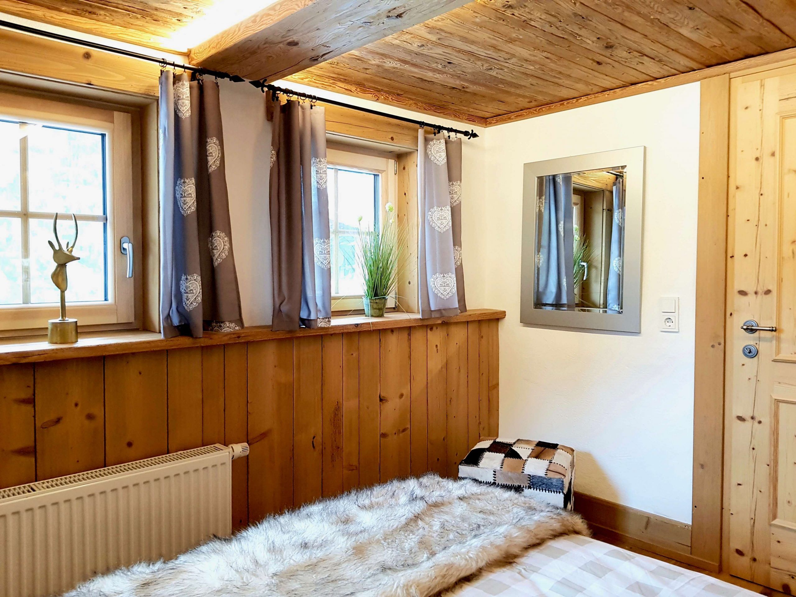 A bedroom with wooden paneling, a radiator, a bed with a fur throw, a window with curtains, and a mirror on the wall.