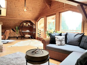 A cozy wooden interior with a sofa, a round table, a bookshelf, and a large window.
