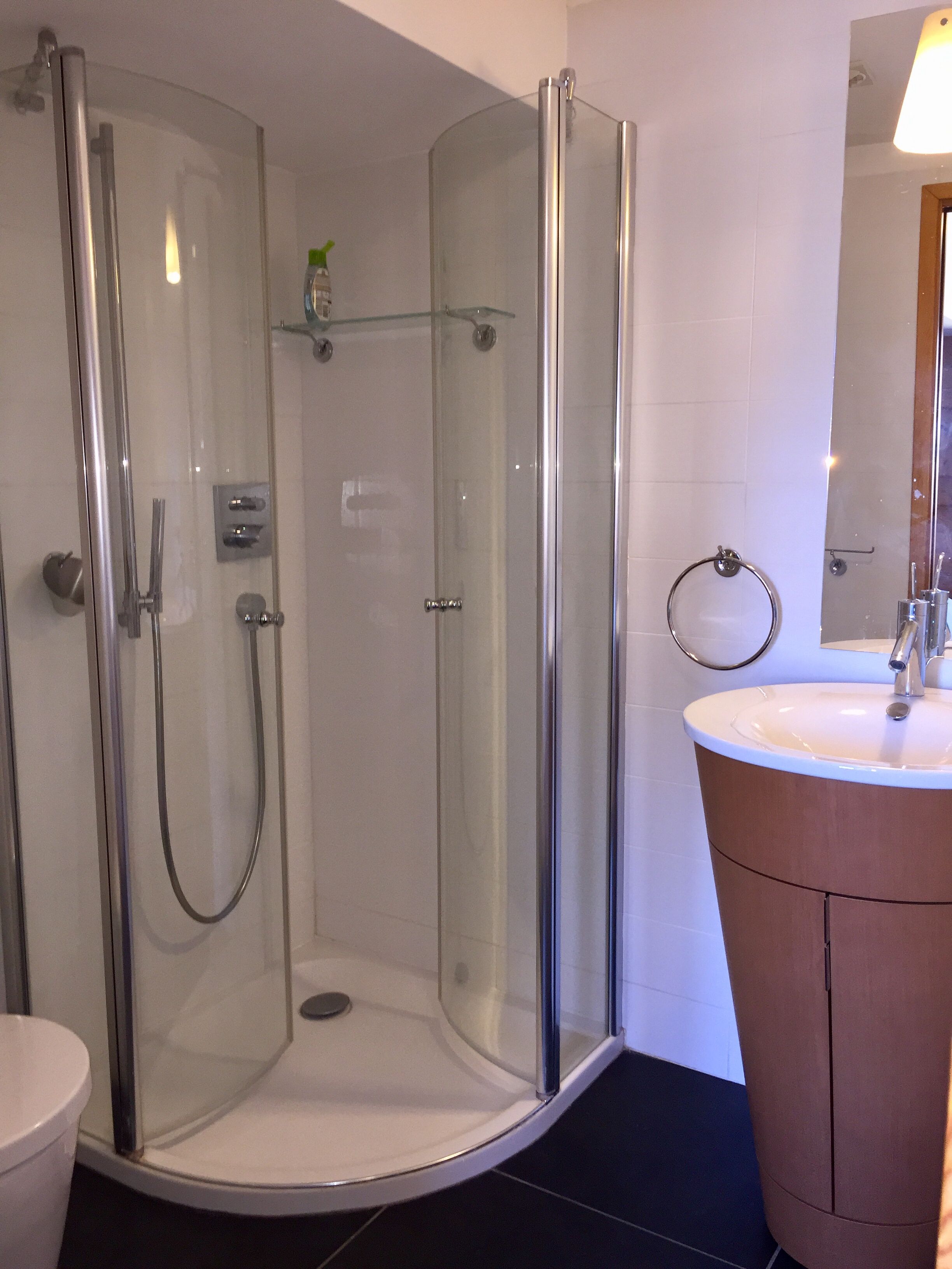 A glass-enclosed shower with a handheld showerhead, a sink with a round mirror, and a small shelf with a bottle.